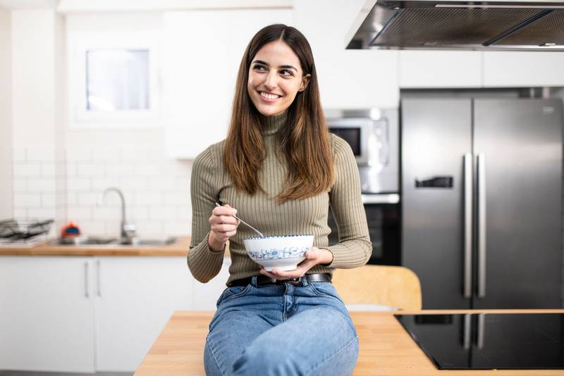 young beautiful woman with a bowl of cereals and a spoon smiling at kitchen - healty, food and female concept