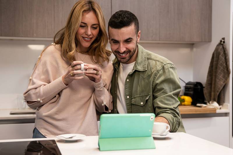 Young adult couple using together a tablet from home in a modern kitchen. Happy joyful man and woman on a video call having a coffee cup.
