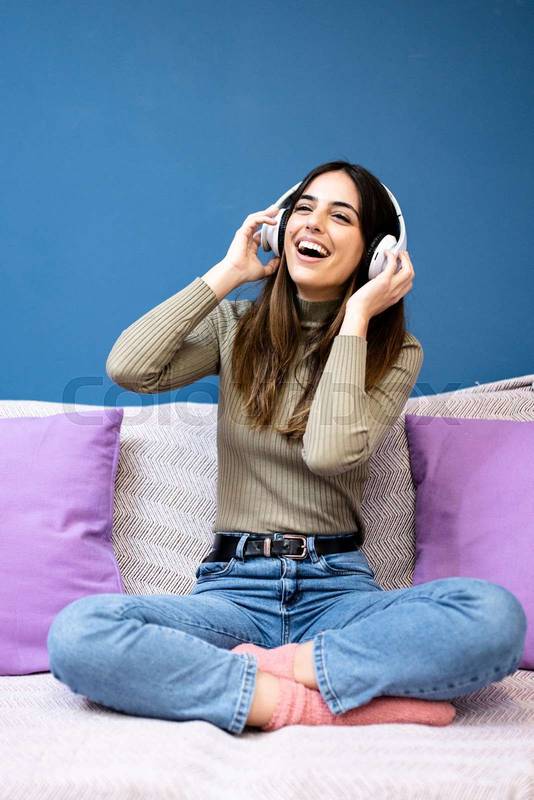 Cheerful young woman listening to music and smiling sitting on couch in livinig room. Happy young girl having fun at home.
