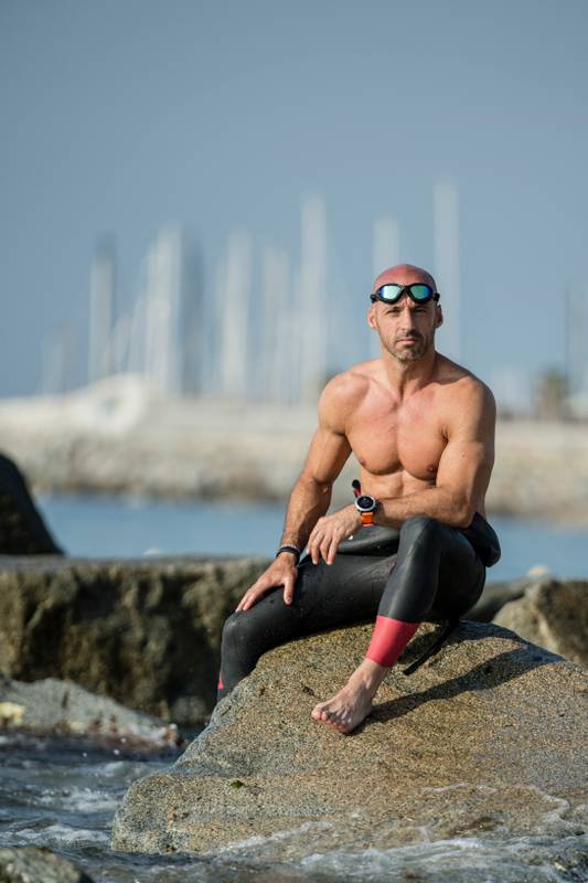Confident fit man sitting on sea rocks with wetsuit and swim glasses. Focused musuclar male sitting by the sea shirtless looking at camera.