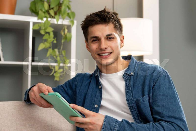 Smiling relaxed male using digital tablet at a modern apartment. Handsome carefree young man working with touchpad sitting on the couch at home looking at camera. 