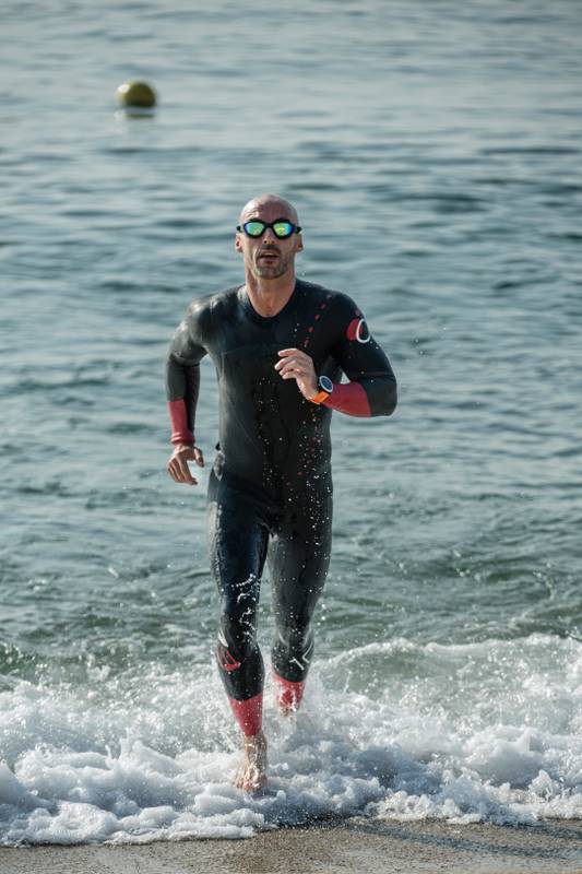 Male swimmer running out of ocean with glasses and wetsuit looking at camera. Male triathlete getting out of the water staring at the camera.