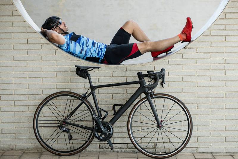Athletic man in sport clothes resting in a wall with a cycle. Fit male cyclist lying next to his cycle wearing helmet and sunglasses.