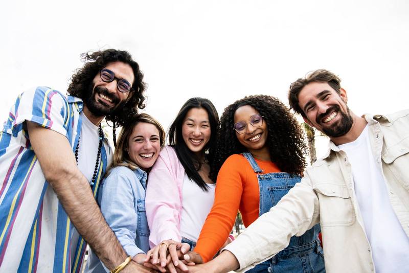 Diverse group of cheerful millennial friends standing together stacking hands in the street looking at camera. Cheerful multiethnic young carefree friends laughing and having fun 