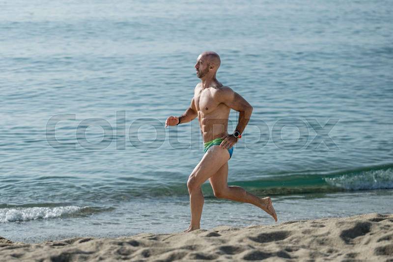 Athletic muscular man in swimwear running next to the sea. Professional male athlete training in speedo at the beach.
