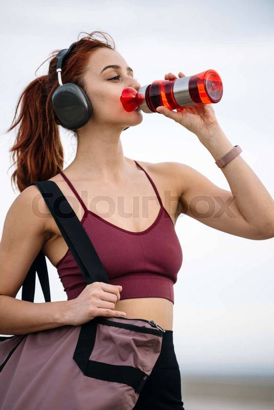 Young fitness female drinking water outside wearing headphones. Athletic woman using a bottle and a gym bag ready for training and workout.