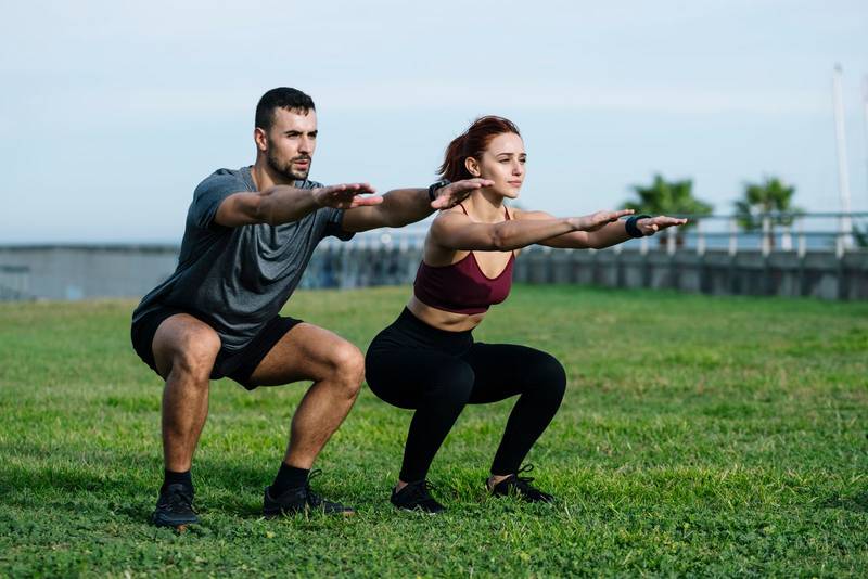 Fit young adult couple exercising outside training for running together in the park. Athletic male and female squatting in the grass in sportswear workout outside. 