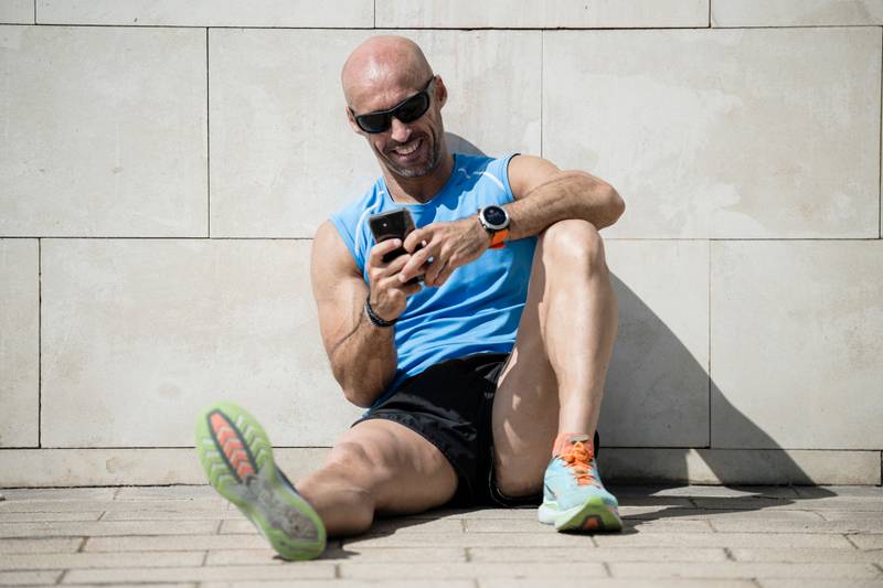 Fit happy man checking his phone sitting on the street floor. Relaxed smiling athletic runner male watching his smartphone resting against a wall in the city.