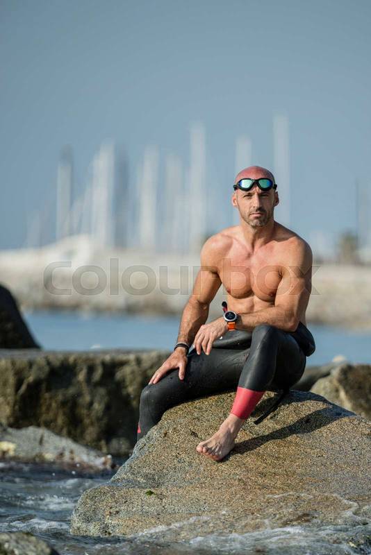 Confident fit man sitting on sea rocks with wetsuit and swim glasses. Focused musuclar male sitting by the sea shirtless looking at camera.