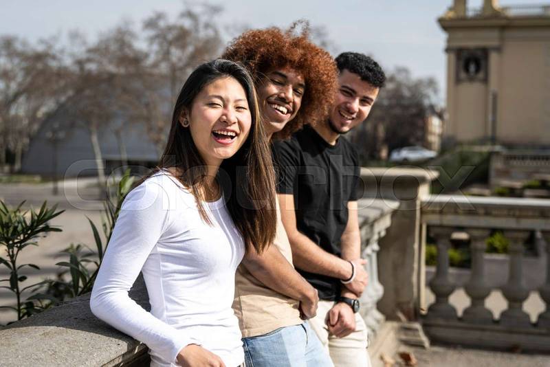 Multiracial group of young laughing people standing on a park looking at the camera. Three diverse happy friends having fun together outside staring at camera.