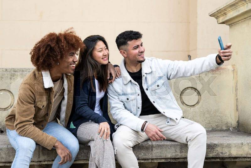 Three young diverse people taking a selfie with a smartphone sitting on a bank.Multiracial group smiling and taking a picture with a phone in a stone park bank.