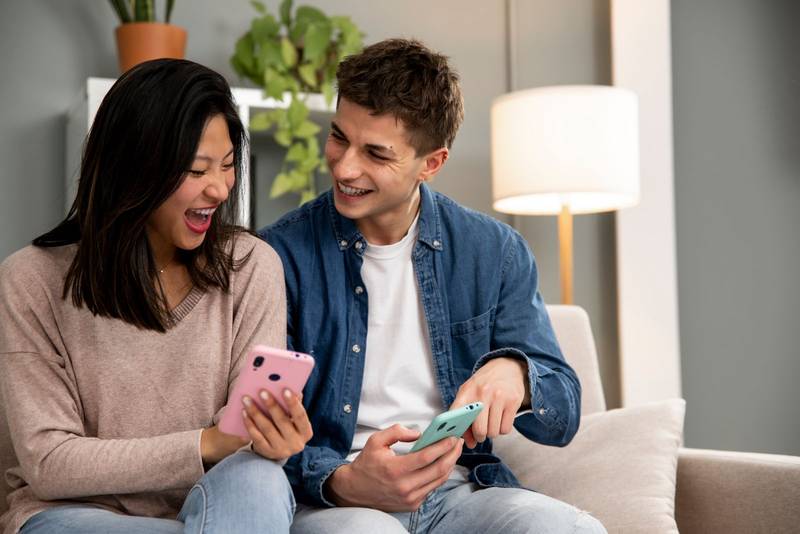 Happy young couple using smartphone relaxing on a couch in their living room at home. Diverse man and woman smiling and having fun using online phones.